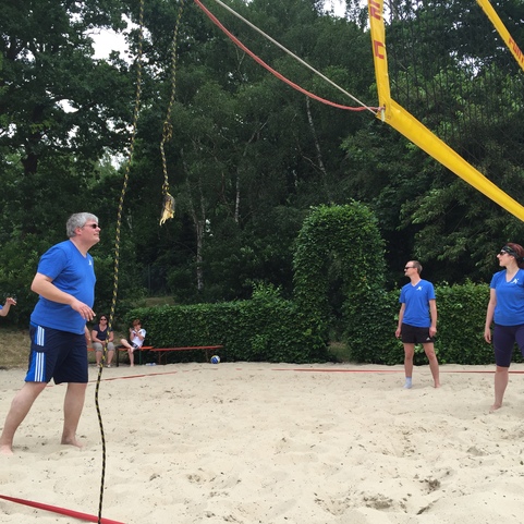 Vier Personen in blauen T-Shirts spielen Beachvolleyball auf einem Sandplatz, umgeben von B&auml;umen.