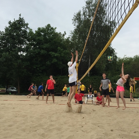 Zwei Spieler springen, um einen Volleyball &uuml;ber das Netz auf einem Sandplatz zu schlagen, umgeben von mehreren anderen Spielern.