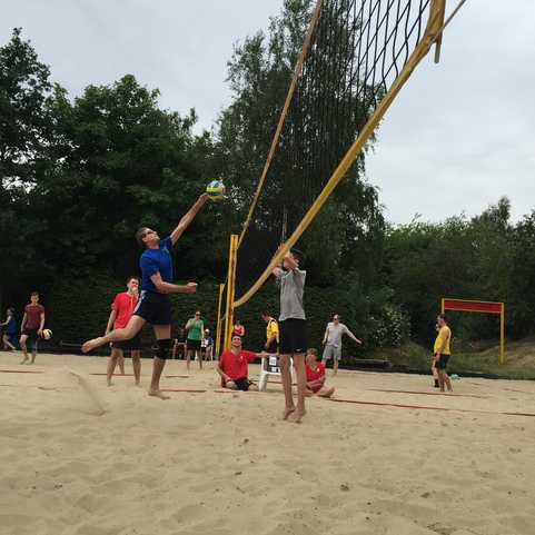 Spieler springen, um einen Ball &uuml;ber ein Netz auf einem Sandplatz zu schlagen, w&auml;hrend andere zuschauen.