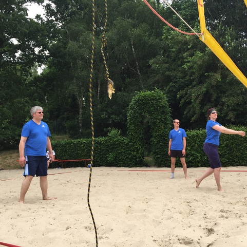 Vier Personen in blauen Shirts spielen Beachvolleyball auf einem Sandplatz, umgeben von B&auml;umen und einem Netz.