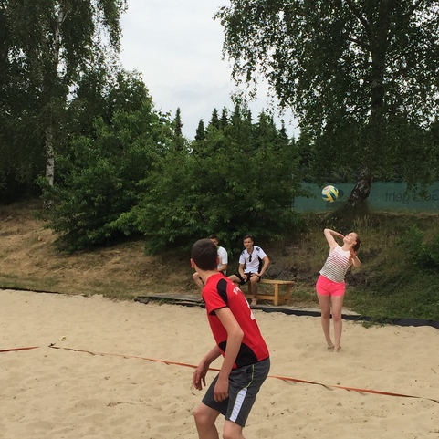 Zwei Kinder spielen im Sand Volleyball; im Hintergrund sitzen zwei Jugendliche auf einer Bank.