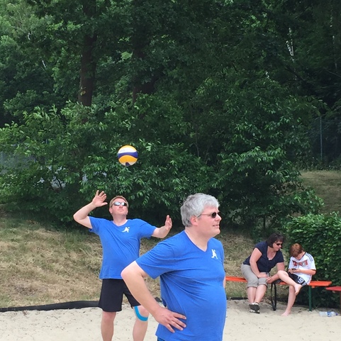 Zwei M&auml;nner in blauen T-Shirts spielen Volleyball am Strand, w&auml;hrend Zuschauer im Hintergrund entspannen.