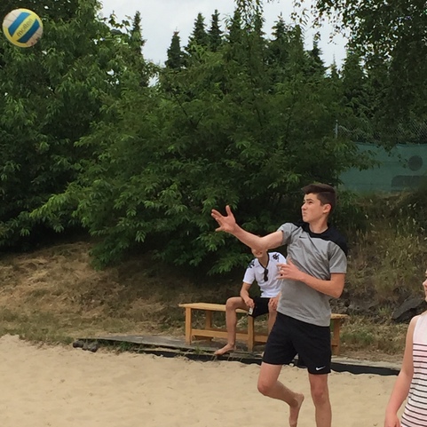 Junge spielt Volleyball am Sandstrand, w&auml;hrend ein Ball in der Luft fliegt und B&auml;ume im Hintergrund sichtbar sind.