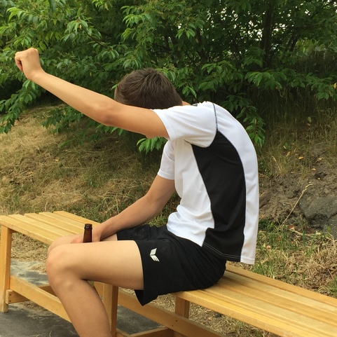 Junger Mann sitzt auf einer Bank, hebt einen Arm und h&auml;lt eine Flasche in der anderen Hand, umgeben von Natur.