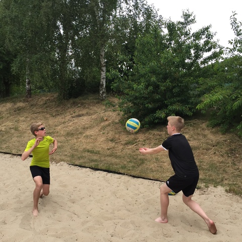 Zwei Jungen spielen im Sand mit einem Volleyball, w&auml;hrend ein dritter Junge im Hintergrund zuschaut.