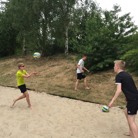 Drei Jungen spielen Beachvolleyball auf einem Sandplatz, w&auml;hrend B&auml;ume im Hintergrund sichtbar sind.