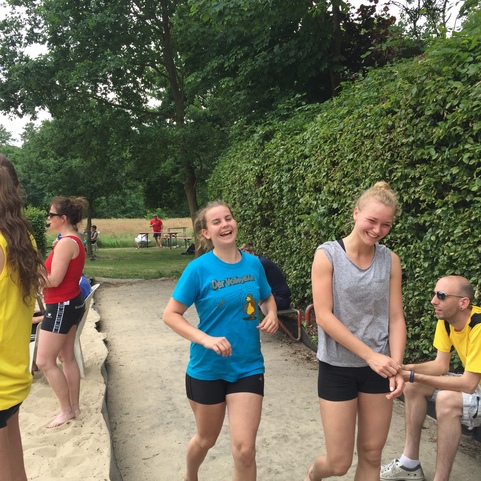 Zwei fr&ouml;hliche M&auml;dchen in Sportkleidung laufen barfu&szlig; auf einem Sandplatz, umgeben von weiteren Personen und B&auml;umen.
