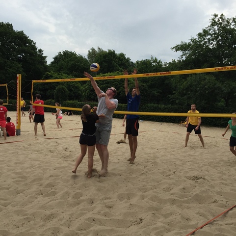 Gruppe von Menschen spielt Volleyball auf einem Sandfeld, w&auml;hrend ein Spieler den Ball &uuml;ber das Netz schl&auml;gt.