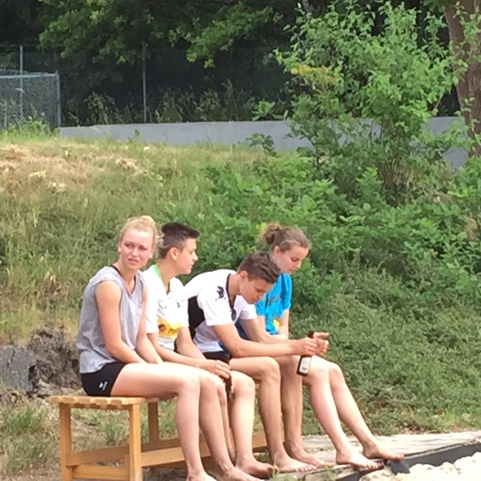 Vier Jugendliche sitzen auf einer Holzbank am Ufer, umgeben von B&auml;umen und Gras. Einige halten Getr&auml;nke in der Hand.