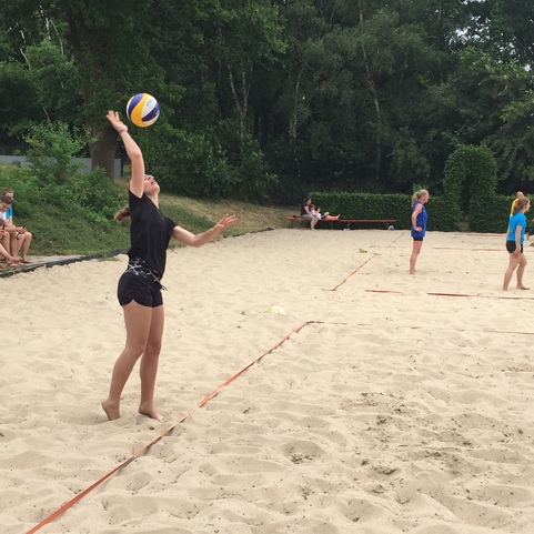 M&auml;dchen serviert einen Volleyball im Sand, w&auml;hrend andere Spielerinnen im Hintergrund warten.