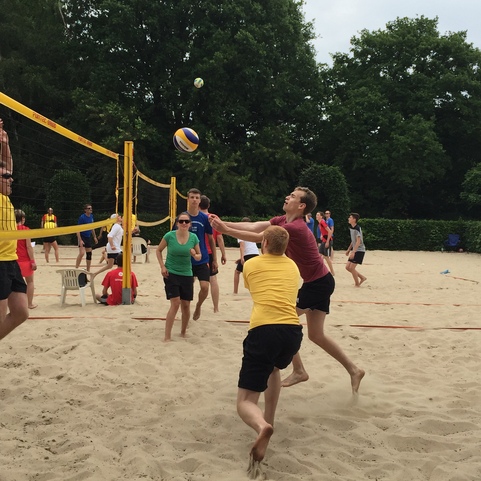 Zwei Spieler im Sand eines Beachvolleyballfeldes springen zum Ball, w&auml;hrend andere zuschauen. B&auml;ume im Hintergrund.