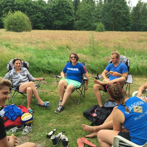 Gruppe junger Erwachsener in Campingst&uuml;hlen sitzt auf einer Wiese, umgeben von B&auml;umen und hoher Graslandschaft.
