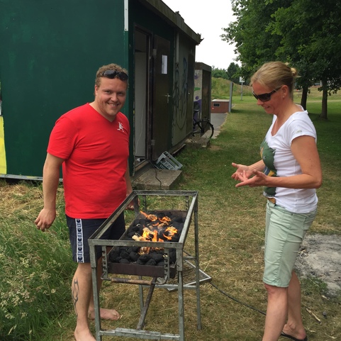 Mann und Frau stehen an einem Grill, der mit brennenden Kohlen gef&uuml;llt ist, vor einem gr&uuml;nen Geb&auml;ude im Freien.