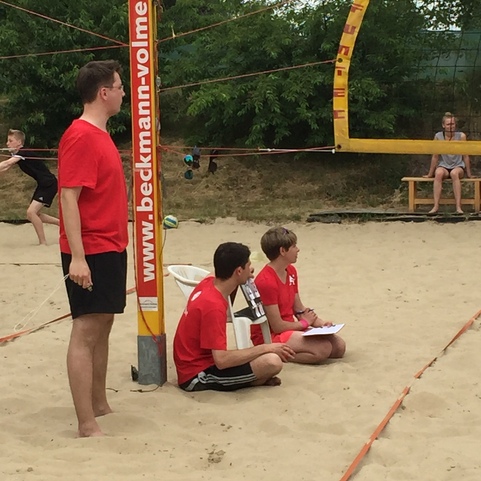 Drei Personen in roter Sportkleidung beobachten ein Beachvolleyballspiel auf einem Sandplatz.