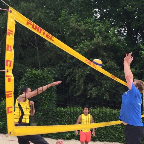 Sportliche Szene im Sandvolleyball: Zwei Spieler springen, um einen Ball &uuml;ber das Netz zu schlagen, w&auml;hrend ein weiterer Spieler zusieht.