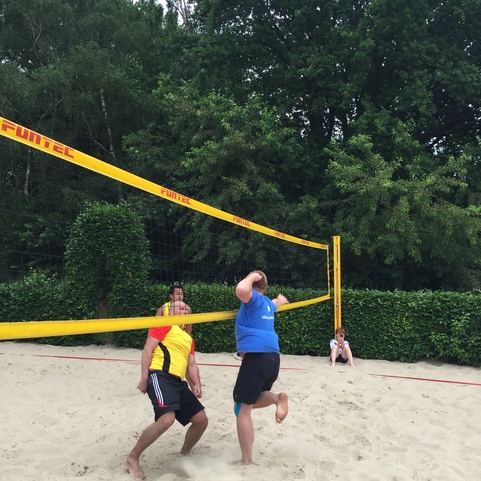 Zwei M&auml;nner spielen Beachvolleyball auf einem Sandplatz, w&auml;hrend eine Person im Hintergrund zusieht.