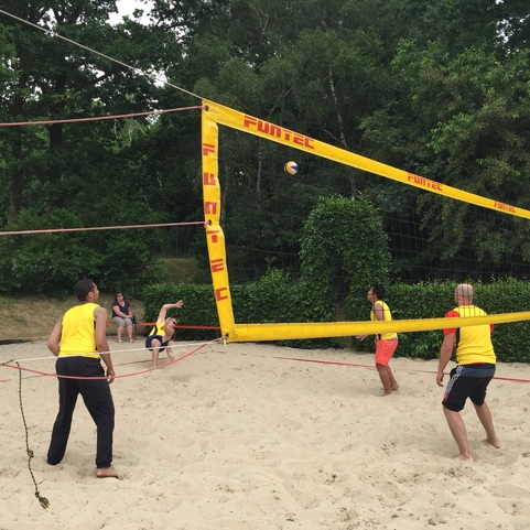 Strandvolleyballspiel mit vier Spielern in gelben Trikots, Ball fliegt &uuml;ber das Netz auf einem Sandplatz.