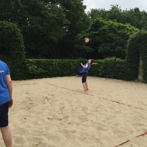 Spieler wirft einen Volleyball in einem Sandfeld, umgeben von gr&uuml;nen Hecken und einem bew&ouml;lkten Himmel.