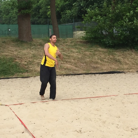 Mann in gelbem Shirt wirft einen Ball &uuml;ber eine rote Linie auf einem Sandplatz, umgeben von B&auml;ume und Wiese.