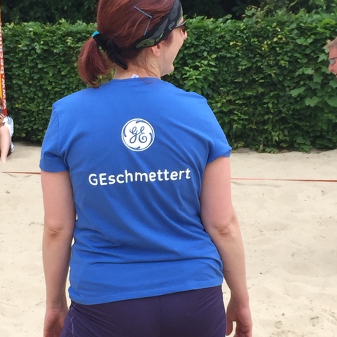 Frau mit rotbraunen Haaren tr&auml;gt ein blaues T-Shirt mit dem Schriftzug "GEs Schmettert" auf einer Sandfl&auml;che.