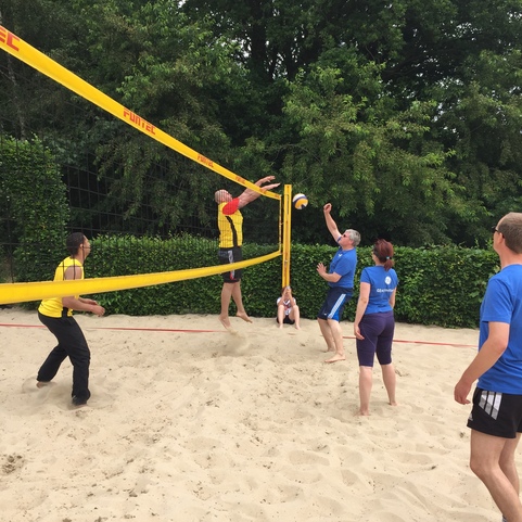 F&uuml;nf Personen spielen Volleyball am Strand; einer springt, um den Ball &uuml;ber das Netz zu schlagen.