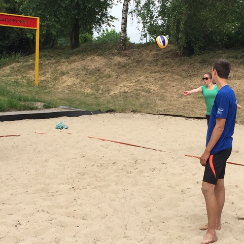 Zwei Personen spielen Volleyball auf einem Sandplatz, w&auml;hrend ein Ball in der Luft schwebt. Im Hintergrund B&auml;ume sichtbar.