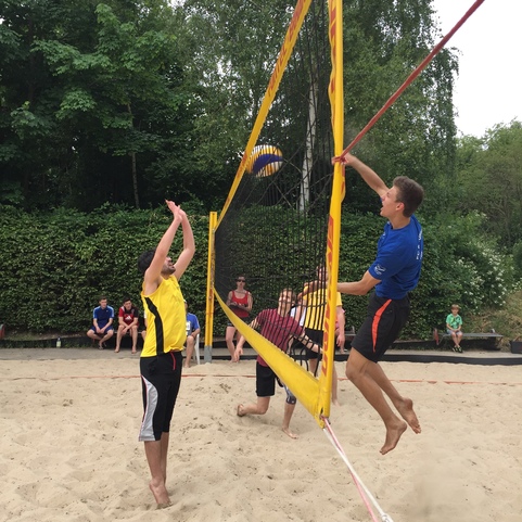 Zwei Spieler im Sand spielen Beachvolleyball, w&auml;hrend Zuschauer im Hintergrund zuschauen. Ein Spieler springt zum Blocken.