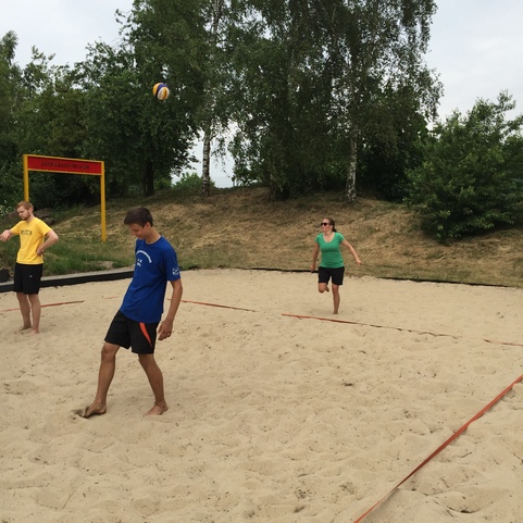 Drei Personen spielen Beachvolleyball auf einem Sandplatz unter bew&ouml;lktem Himmel, im Hintergrund B&auml;ume.