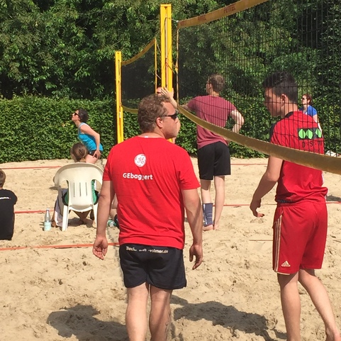 Gruppenszene im Sand mit Volleyballspieler, Netzanlage und Zuschauern in einem Freiluftbereich.