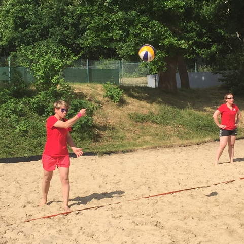 Eine Frau in rotem Shirt schl&auml;gt einen Beachvolleyball &uuml;ber das Netz, w&auml;hrend eine weitere Frau im Hintergrund zusieht.