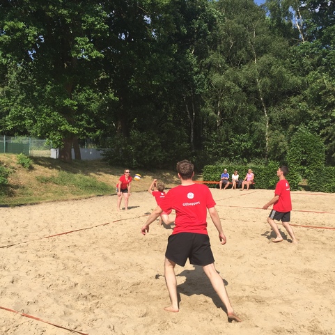 Vier Personen in roten T-Shirts spielen Beachvolleyball auf sandigem Gel&auml;nde, im Hintergrund Zuschauer auf B&auml;nken.
