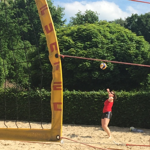 Spielerin in rotem Shirt schl&auml;gt einen Volleyball &uuml;ber das Netz auf einem Sandplatz im Freien.