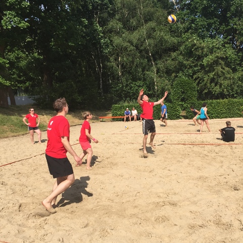 Sandvolleyballspiel mit mehreren Spielern in roten Shirts auf einem Freiluftplatz, umgeben von B&auml;umen.