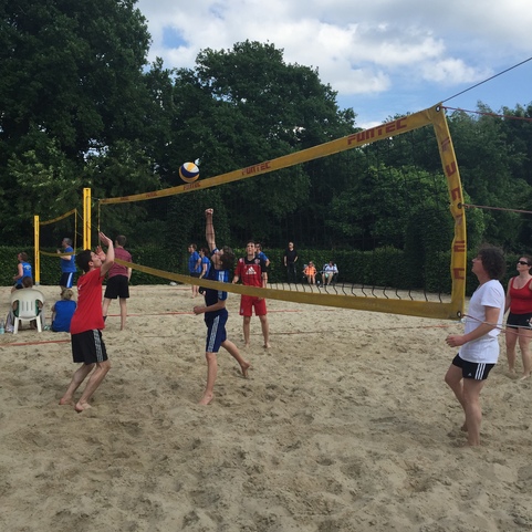 Gruppe von sechs Personen spielt Beachvolleyball auf einem Sandplatz unter einem bew&ouml;lkten Himmel.