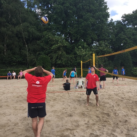 Gruppe von Jugendlichen spielt Volleyball auf einem Sandplatz, im Hintergrund B&auml;ume und ein Volleyballnetz.