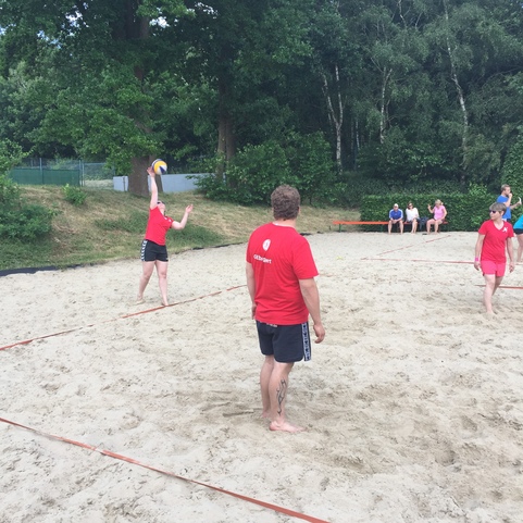 Zwei Personen in roten T-Shirts spielen Beachvolleyball auf einem Sandfeld, im Hintergrund Zuschauer und B&auml;ume.