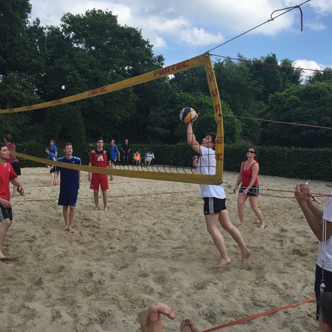 Spielende Menschen beim Beachvolleyball, eine Person springt zum Ball unter dem Netz, umgeben von Zuschauern im Sand.