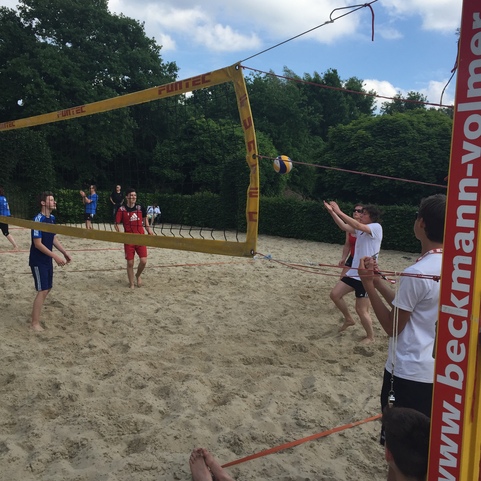 Spieler in blauen und roten Trikots spielen Beachvolleyball auf einem Sandplatz unter einem bew&ouml;lkten Himmel.