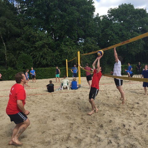 Zwei Spieler heben im Sand einen Volleyball &uuml;ber das Netz, w&auml;hrend andere an einem Sandplatz zuschauen.