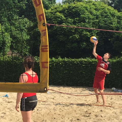 Zwei Personen spielen Beachvolleyball; ein Spieler hebt den Ball zum Aufschlag, w&auml;hrend die andere zusieht.