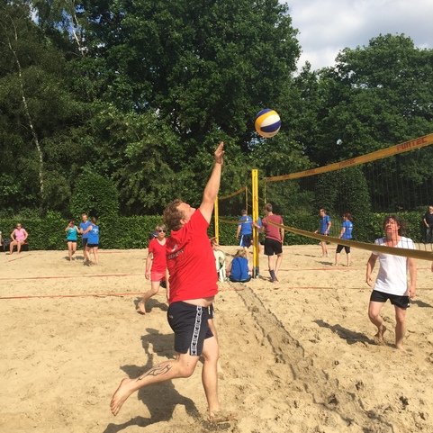 Spieler springt im Sand, um einen Volleyball in ein Beachvolleyballnetz zu schlagen, w&auml;hrend andere Spieler im Hintergrund stehen.