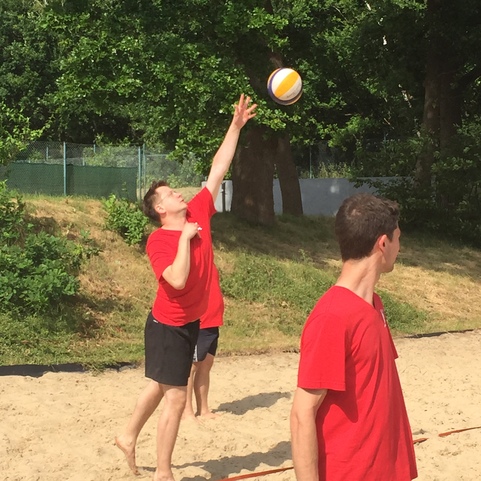 Zwei Jungen in roten T-Shirts spielen Beachvolleyball. Einer springt, um den Ball zu schlagen. Sand und B&auml;ume im Hintergrund.
