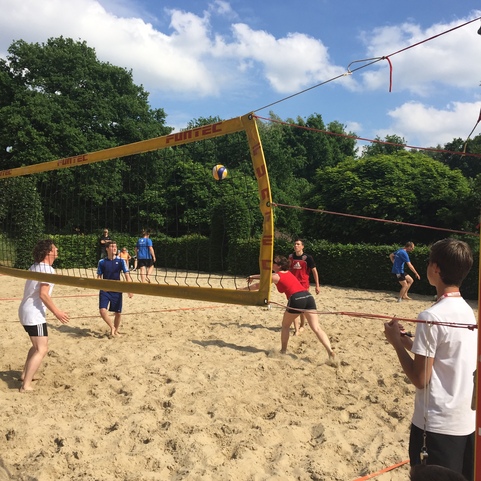 Gruppe von Jugendlichen spielt Volleyball auf einem Sandplatz, umgeben von B&auml;umen und blauem Himmel.