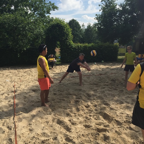 Drei Personen spielen Beachvolleyball auf einem Sandplatz im Freien, umgeben von B&auml;umen und Sonnenschein.