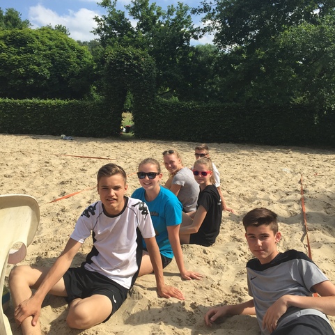 Gruppe von f&uuml;nf Jugendlichen sitzt entspannt im Sand, umgeben von B&auml;umen und blauen Himmel.