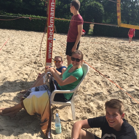 Gruppe von jungen Menschen sitzt entspannt auf einem Volleyballfeld aus Sand, umgeben von B&auml;umen und Netzen.