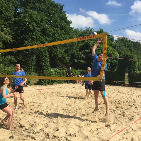 Spieler beim Beachvolleyball in kurzer Sportbekleidung heben einen Ball &uuml;ber das Netz auf einem Sandplatz.