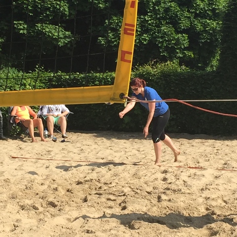 Frau in blauer Sportbekleidung spielt Volleyball am Sandplatz unter einem sonnigen Himmel. Zuschauer sitzen im Hintergrund.