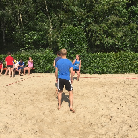 Zwei Jugendliche spielen im Sand, w&auml;hrend Zuschauer auf einer Bank im Hintergrund entspannen. Gr&uuml;ne B&auml;ume umgeben den Bereich.