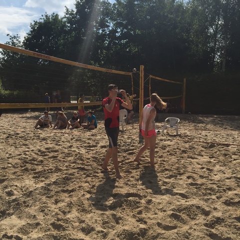 Zwei Jugendliche gehen &uuml;ber einen Sandvolleyballplatz, im Hintergrund sitzen weitere Personen auf St&uuml;hlen.